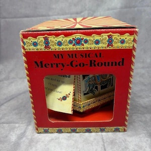 May include: A red and yellow cardboard box for a musical merry-go-round toy. The box features a cutout window displaying a carousel inside, with text reading My Musical Merry-Go-Round and Please wind me up. I'll spin, and play a tune!