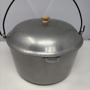 May include: A round, brushed aluminum cooking pot with a matching lid. The lid features a small, light-colored wooden knob handle. A thin, dark metal wire bail handle is attached to the sides of the pot, allowing for easy carrying and hanging.