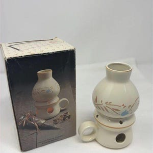May include: A cream-colored ceramic oil burner shaped like a miniature lantern with a side handle and a circular opening at the base. The burner features a painted floral pattern in blue and brown. It is displayed next to its original cardboard box.