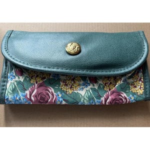 May include: A teal-colored clutch with a floral pattern. The clutch features a flap closure with a gold-colored button. The floral pattern includes roses, blue flowers, and yellow flowers on a white background.