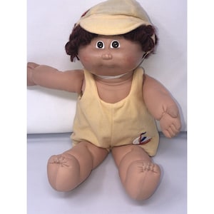 May include: A vintage doll with brown hair, wearing a yellow swimsuit and a matching cap. The doll has a round face with rosy cheeks and large, black eyes. The swimsuit features a small embroidered design. The doll is sitting with its arms outstretched.