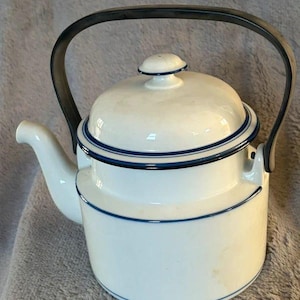 May include: A white enamel teapot featuring a cylindrical body, a curved spout, and a matching lid with a small knob. The teapot is accented with thin, dark blue horizontal stripes along the lid rim and the body. It has a large, dark metal handle.