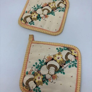 May include: Two square fabric pot holders with a cream background, featuring a printed pattern of white mushrooms, yellow flowers, and green leaves. The edges are trimmed with a yellow and orange polka dot fabric and each has a small hanging loop.