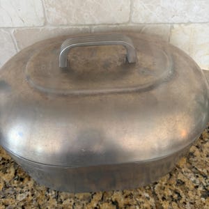May include: An oval-shaped metal roasting pan with a matching lid. The exterior has a brushed silver finish with visible signs of wear and patina. A rectangular handle with three horizontal ridges is centered on the top of the lid.