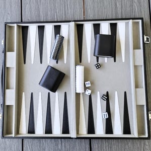 May include: Open backgammon set with black and white playing pieces, dice, and cups. The game board is black with white and black triangles. The set includes dice cups and dice. The logo "JouZ" is in the bottom left corner.