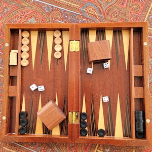 May include: A wooden backgammon set, open and ready to play. The set features a dark wood finish, with light and dark-colored triangles. White and black game pieces are arranged on the board, along with dice and wooden dice cups. The word "jouz" is visible.