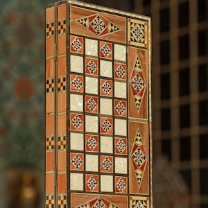Op de afbeelding: Een houten backgammonbord met ingewikkelde inlegontwerpen. Het bord heeft een dambordpatroon met rode, zwarte en witte vierkanten, omlijst door geometrische patronen in bruin, rood en geel. Het bord staat rechtop.