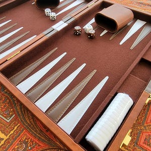 May include: A brown leather backgammon set with white and brown playing pieces. The set is open, revealing the playing surface, dice, and dice cups. The game board is brown with white and brown triangles.