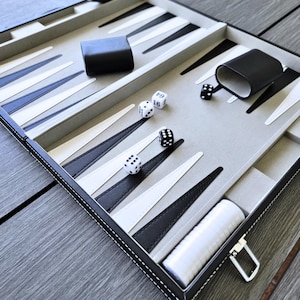 May include: A backgammon set with a gray playing surface, black and white triangles, and dice. The set includes a black case, dice cups, and a white container for game pieces. The board is framed in black with white stitching.