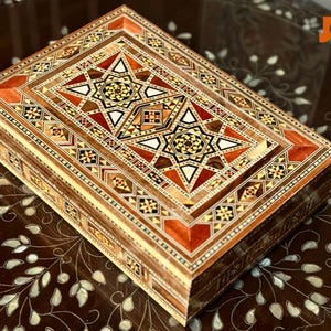 May include: A rectangular wooden box with detailed inlaid geometric patterns in brown, red, and cream. The lid features a starburst design. Likely used for storage or display, the box is a decorative item.