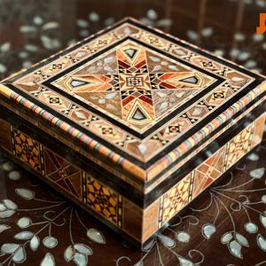 Könnte beinhalten: Quadratische Holzbox mit aufwendigen eingelegten geometrischen Mustern in Braun-, Orange- und Schwarztönen. Die Box hat ein zentrales Sternmuster und steht auf einer dunklen, gemusterten Oberfläche. Dekorative Aufbewahrungsbox.
