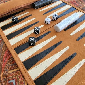 May include: A brown backgammon board with black, white, and brown triangular points. The board is open, with dice and dice cups visible. The board is made of suede-like material and has a zippered compartment.
