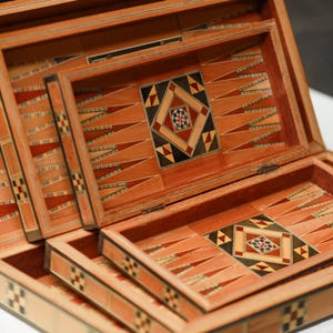 May include: Wooden backgammon set with intricate inlaid geometric designs in shades of brown, red, black, and cream. The game board features triangular points and a central decorative square. The set is open, revealing the playing surface and storage compartments.