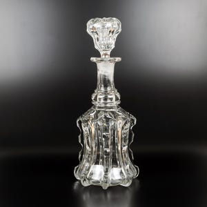 May include: A clear glass decanter with a decorative stopper. The decanter has a bulbous base and a long neck, with a detailed, textured design. The stopper is also clear and has a flower-like shape. The background is a gradient of black and grey.