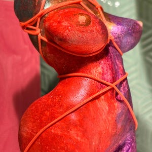 Könnte beinhalten: Eine Torsoskulptur mit einer lebendigen Mischung aus Rot-, Orange- und Violetttönen mit strukturierter Oberfläche. Die Figur ist mit einer dünnen, leuchtend neonorangen Kordel umwickelt, die in einem dekorativen Muster über Brust, Taille und Hüften gebunden ist.