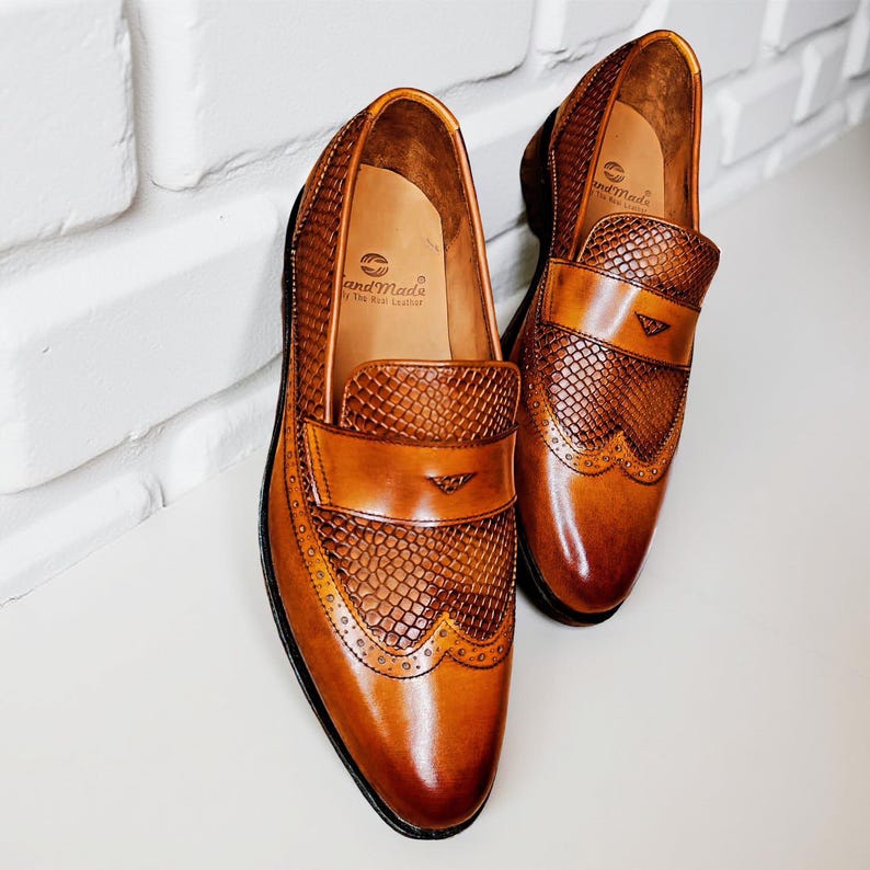 Pu&ograve; includere: Un paio di mocassini in pelle marrone con tomaia strutturata e dettagli brogue. Le scarpe hanno una finitura lucida e un piccolo stemma sul cinturino. L'interno della scarpa &egrave; visibile, con la scritta "and Made" stampata sulla soletta.