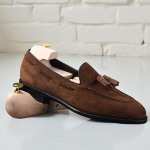 May include: A brown suede tassel loafer shoe featuring a black sole and a light-colored wooden shoe tree inserted inside. The shoe has a rounded toe, visible stitching along the vamp, and a small decorative tassel detail on the top surface.