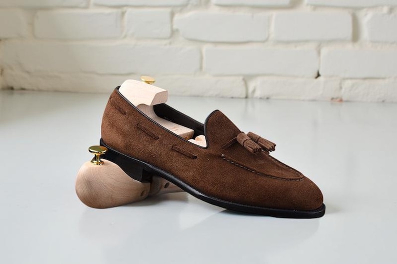 May include: A brown suede tassel loafer shoe featuring a black sole and a light-colored wooden shoe tree inserted inside. The shoe has a rounded toe, visible stitching along the vamp, and a small decorative tassel detail on the top surface.