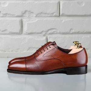May include: A pair of cognac brown leather oxford shoes featuring a cap toe design, dark brown laces, and a low stacked heel. The shoes are equipped with light-coloured wooden shoe trees with gold-toned metal hardware inserted inside.