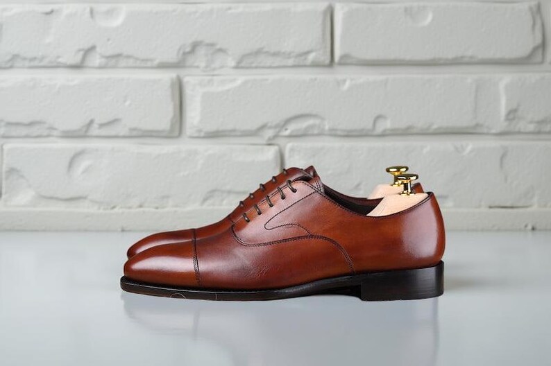 May include: A pair of cognac brown leather oxford shoes featuring a cap toe design, dark brown laces, and a low stacked heel. The shoes are equipped with light-coloured wooden shoe trees with gold-toned metal hardware inserted inside.