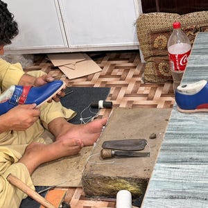 May include: A pair of blue and red monk strap shoes with silver buckles. One shoe is being worked on by hand, while the other sits on a table. The shoes feature a smooth finish, a rounded toe, and a distinct color-blocked design with red straps.