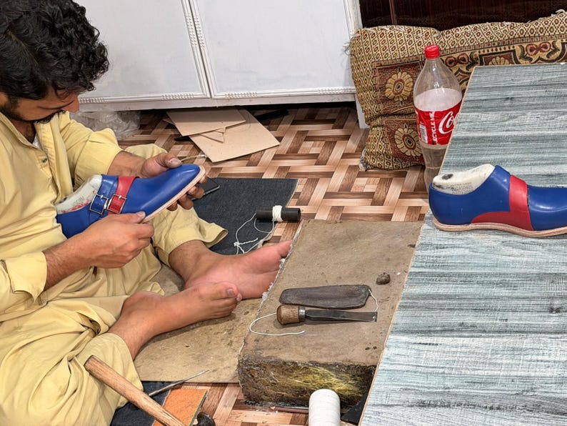 May include: A pair of blue and red monk strap shoes with silver buckles. One shoe is being worked on by hand, while the other sits on a table. The shoes feature a smooth finish, a rounded toe, and a distinct color-blocked design with red straps.