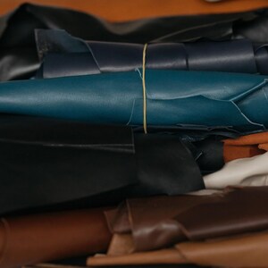 May include: A collection of rolled leather sheets in various colors including black, dark blue, teal, cream, and shades of brown. The rolls are secured with thin rubber bands, displaying the smooth texture and flexible nature of the material.