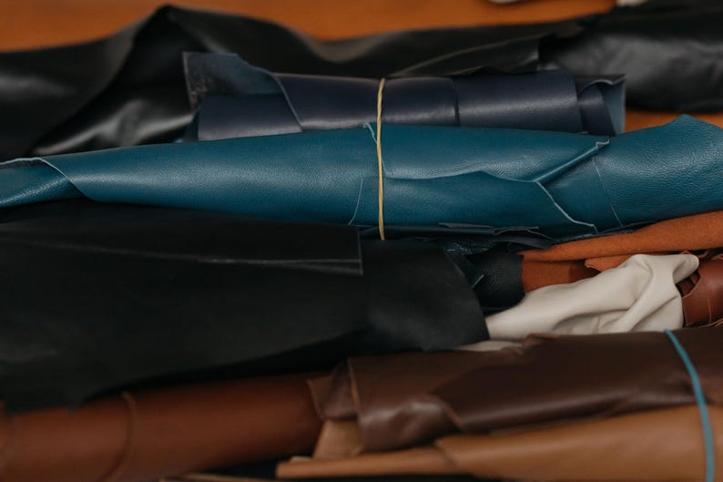 May include: A collection of rolled leather sheets in various colors including black, dark blue, teal, cream, and shades of brown. The rolls are secured with thin rubber bands, displaying the smooth texture and flexible nature of the material.