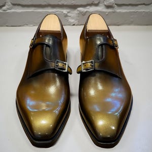 Pode incluir: Um par de sapatos de fivela polidos com um acabamento degradê em verde-azeitona e castanho-escuro. Cada sapato possui um fecho de fivela de metal prateado e uma forma de madeira clara inserida na abertura para manter a sua forma.