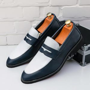 May include: A pair of navy and white leather loafers. The shoes feature a white vamp and strap detail, with a navy blue body. The soles are black with a red edge. Wooden shoe trees are inside the shoes.