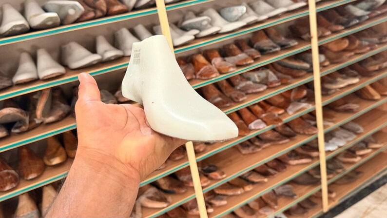 May include: A white plastic shoe last with a smooth, contoured shape designed for footwear manufacturing. The object features a pointed toe and a raised heel section, with faint text markings visible on the side of the ankle area.