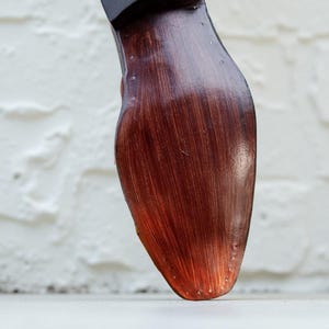 Op de afbeelding: Close-up van een bruine leren schoenzool met een donkere hiel. De zool heeft een roodbruine tint met zichtbare nerf en stiksels. De schoen staat tegen een witte achtergrond en een getextureerde witte muur.