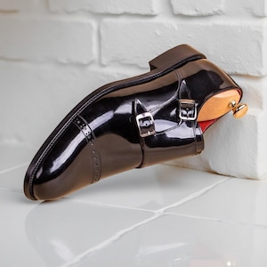 May include: A black patent leather double monk strap shoe with a cap toe featuring decorative brogue perforations. The shoe includes two silver-toned metal buckles and a wooden shoe tree inserted inside with a gold-toned knob at the heel.