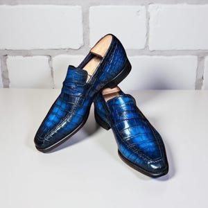 以下が含まれることがあります： クロコダイル柄のブルーレザーローファーのペア。光沢のある仕上がりと、クラシックなスリッポンデザインです。白い表面に展示されており、背景には白いレンガの壁があります。