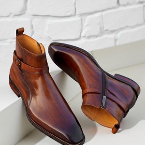 May include: A pair of brown leather ankle boots with a polished finish. The boots feature a side zipper, a buckle strap, and a square toe. The leather has a gradient effect, transitioning from light to dark brown.