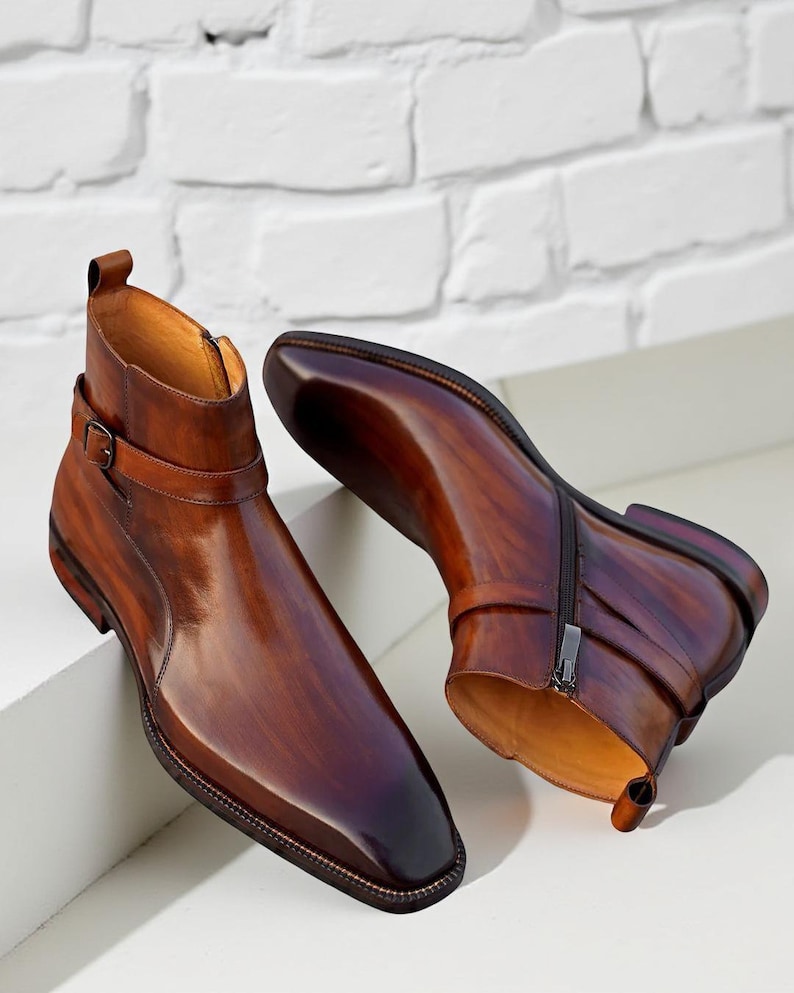 May include: A pair of brown leather ankle boots with a polished finish. The boots feature a side zipper, a buckle strap, and a square toe. The leather has a gradient effect, transitioning from light to dark brown.