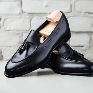 May include: A pair of black leather loafers with tassel details. The shoes are displayed with wooden shoe trees inside, and are set against a white brick wall background. The loafers have a classic, polished appearance.