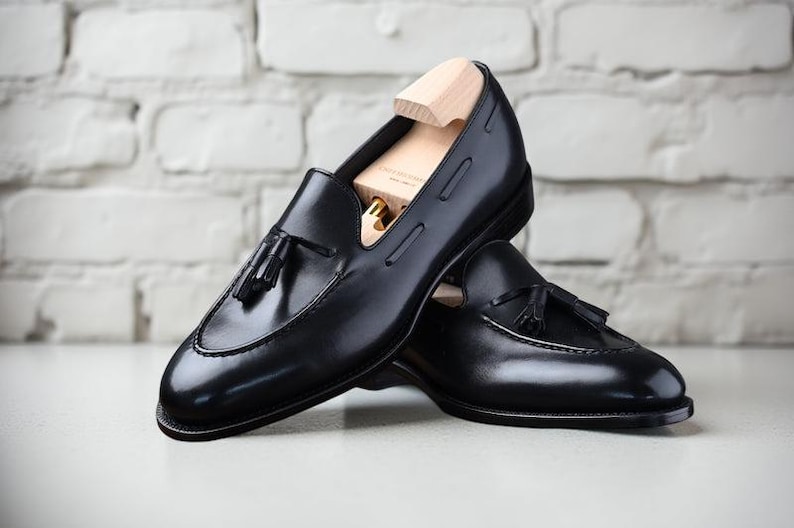 May include: A pair of black leather loafers with tassel details. The shoes are displayed with wooden shoe trees inside, and are set against a white brick wall background. The loafers have a classic, polished appearance.