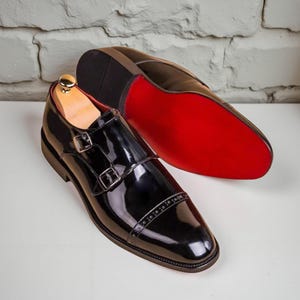 May include: A pair of black patent leather double monk strap shoes with silver-toned buckles and a cap toe featuring decorative brogue detailing. One shoe is positioned upright, while the other is placed behind it, revealing a bright red sole.