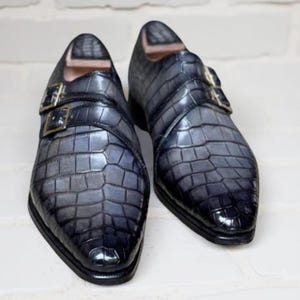 K&ouml;nnte beinhalten: Ein Paar blau-graue Lederschuhe mit Krokodilleder-Textur. Die Schuhe haben ein Doppel-M&ouml;nch-Riemen-Design mit goldfarbenen Schnallen. Die Schuhe werden auf einem wei&szlig;en Ziegelhintergrund pr&auml;sentiert.