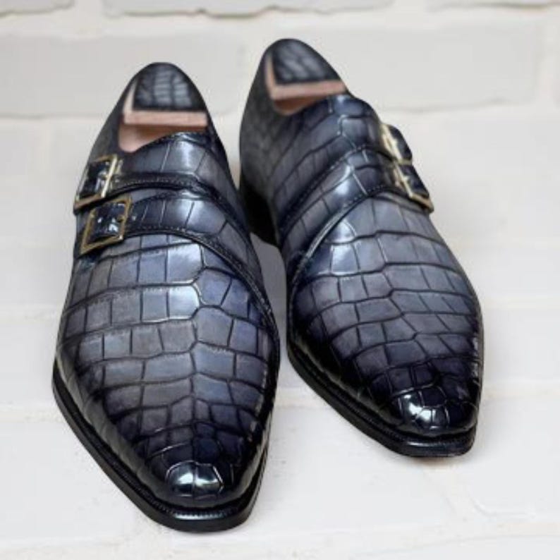 K&ouml;nnte beinhalten: Ein Paar blau-graue Lederschuhe mit Krokodilleder-Textur. Die Schuhe haben ein Doppel-M&ouml;nch-Riemen-Design mit goldfarbenen Schnallen. Die Schuhe werden auf einem wei&szlig;en Ziegelhintergrund pr&auml;sentiert.