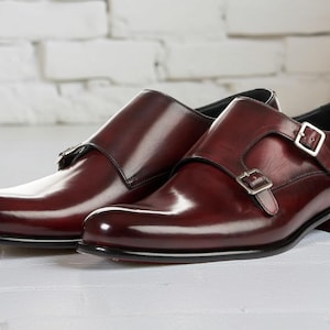 Pode incluir: Um par de sapatos de couro polido em tom bordô escuro com fivela dupla, apresentando fivelas de metal prateado nas tiras laterais, bico arredondado e salto baixo preto. O couro tem um acabamento liso e brilhante com sombreamento tonal.