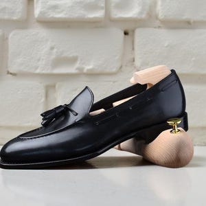 May include: A black leather loafer with a tassel detail, displayed with a wooden shoe tree. The shoe has a sleek design and is set against a white brick wall background.