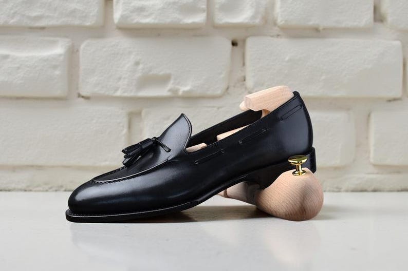 May include: A black leather loafer with a tassel detail, displayed with a wooden shoe tree. The shoe has a sleek design and is set against a white brick wall background.