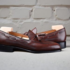 May include: A pair of brown leather loafers with a classic design. The shoes feature a tassel detail and a low heel. The loafers are displayed on a light-coloured surface against a white brick wall.