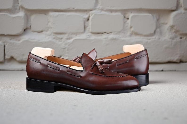 May include: A pair of brown leather loafers with a classic design. The shoes feature a tassel detail and a low heel. The loafers are displayed on a light-coloured surface against a white brick wall.