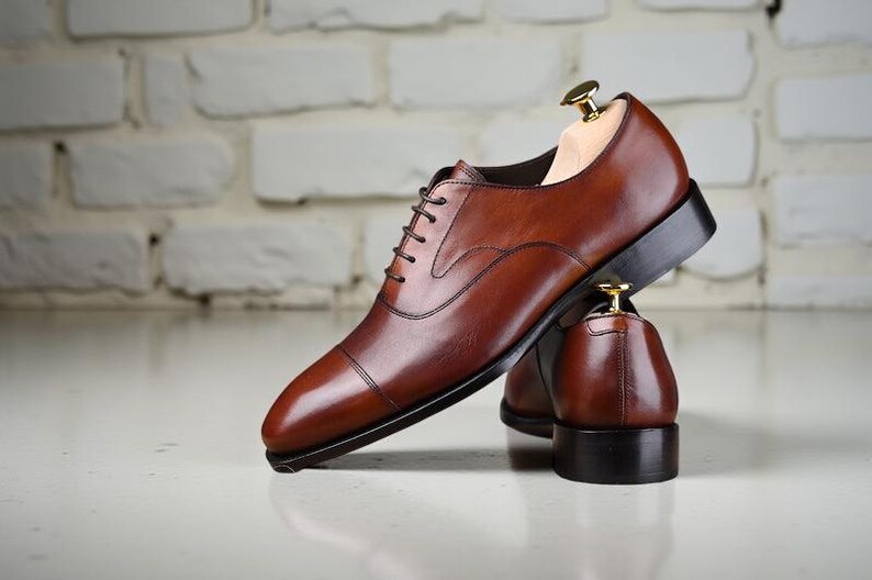 May include: A pair of brown leather oxford shoes with a cap toe design, dark brown laces, and a black sole. Each shoe features a light-coloured wooden shoe tree inserted inside to maintain the shape of the leather upper and the heel structure.