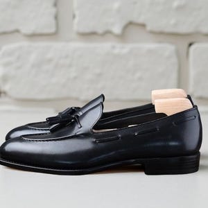 May include: A pair of black leather loafers with tassels. The shoes have a glossy finish and are displayed with wooden shoe trees inside. The background is a white brick wall.