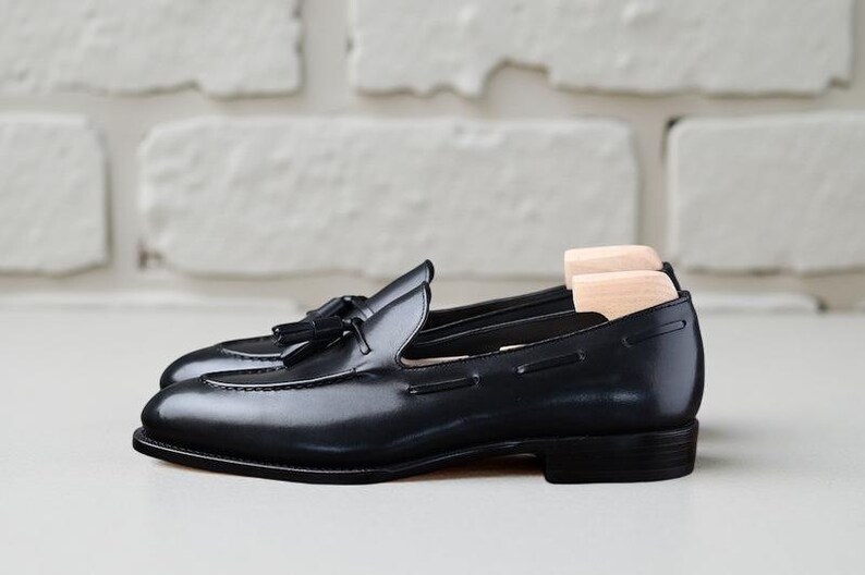 May include: A pair of black leather loafers with tassels. The shoes have a glossy finish and are displayed with wooden shoe trees inside. The background is a white brick wall.