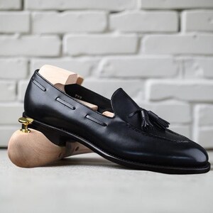 May include: Black leather loafer with a tassel detail, resting on a wooden shoe tree. The shoe has a sleek design and is set against a white brick wall background.